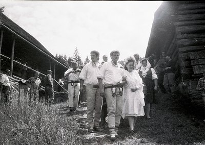 Rural gathering in mid-20th century, likely 1950s–1960s. Group of people in casual attire—men in trousers/suspenders, women i...