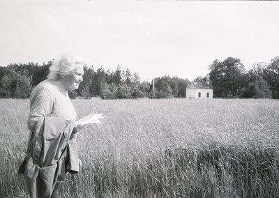 Black-and-white portrait of an elderly woman in a rural field, holding papers while wearing a sweater and a crossbody bag. Ab...