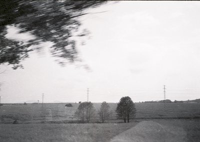 Mid-20th century rural landscape with motion blur from moving vehicle. Open fields, sparse trees, and utility poles dominate ...