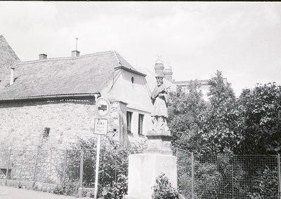Black-and-white street scene featuring a stone statue of a robed figure holding a scroll, likely a historical or religious mo...
