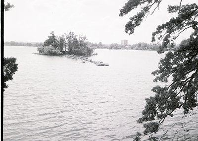 Black-and-white lakeside scene featuring a small island with a single structure and dock. Overcast skies and calm waters domi...