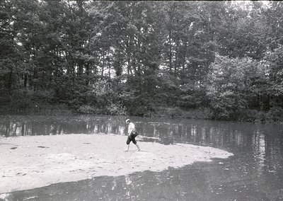 A lone individual wades through shallow, reflective water bordered by dense forest foliage. Black-and-white vintage photograp...