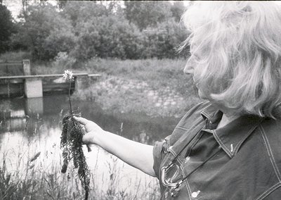 Mid-century woman in outdoor setting examines aquatic plant near waterway, likely for botanical study or environmental observ...