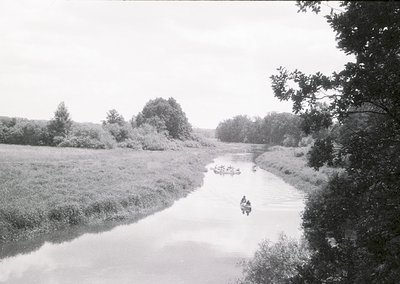 Vintage black-and-white scene of a narrow river flanked by dense reeds, with two small boats carrying people. One boat appear...