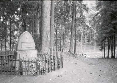 Monumental stone obelisk with engraved plaque in a wooded forest path, surrounded by a rustic wooden fence. Snow covers groun...