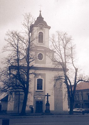 Neoclassical church with tall bell tower featuring a clock and cross, flanked by bare trees. Architectural details include ar...