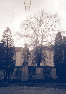 Statue of a robed figure on pedestal in courtyard, flanked by trees and a building with Cyrillic lettering. Likely Eastern Eu...