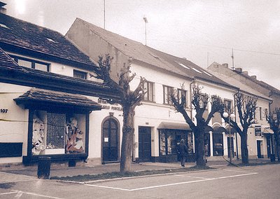 Mid-20th century European street corner with two-story brick buildings. Left: "Pizzeria" sign with vintage mural of figures i...