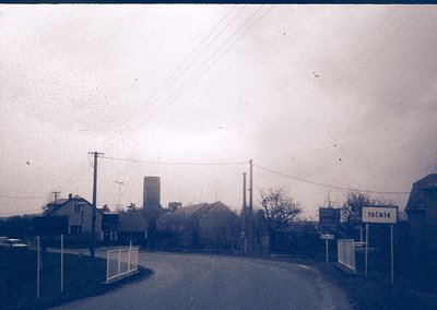 Vintage black-and-white street scene featuring a small industrial area with a tall brick chimney and residential houses. A si...