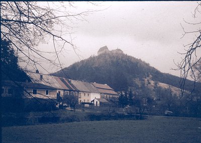 Vintage black-and-white village scene with clustered brick houses under a rocky hilltop fortress. Bare winter trees frame the...