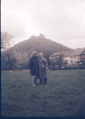 Vintage black-and-white photo of two women posing in a grassy field with a rocky hillock and rustic houses in background. Mid...