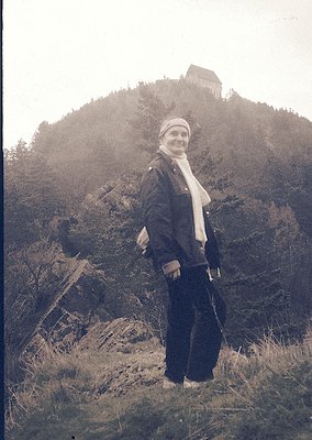 Vintage sepia-toned portrait of a woman in outdoor attire—dark jacket, patterned scarf, and trousers—standing on a rocky hill...