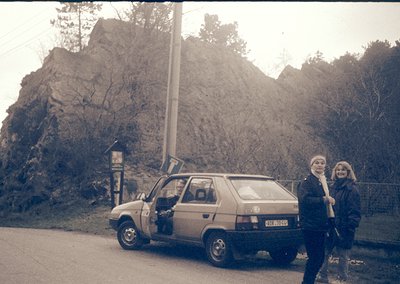 Vintage car parked beside rugged rock formations with two women in 1970s-style clothing. The car’s license plate suggests Eas...