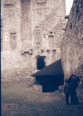 Medieval stone courtyard with weathered walls and narrow alleyway. A small, sloped-roof structure (likely a guardhouse or sto...