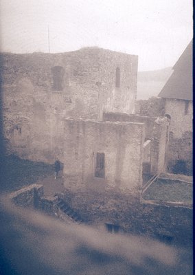 Mid-20th century black-and-white photo of weathered stone ruins with crumbling walls and arched windows. A lone figure stands...