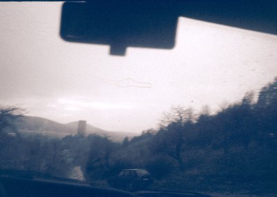 Vintage rearview shot of a misty, rural road with sparse traffic and distant hills. Blurred greenery and a lone car suggest a...