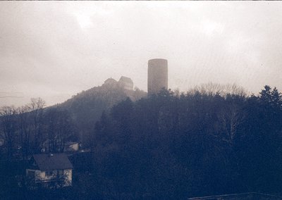Vintage sepia-toned photo of a medieval hilltop fortress with a cylindrical tower, surrounded by sparse winter trees. Residen...