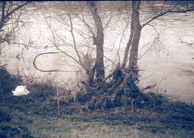 Vintage sepia-toned photo of a rural riverside scene with leafless trees and shallow water. A faint, partially submerged obje...