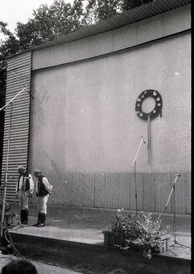 Indoor stage with concrete wall backdrop featuring a wreath emblem. Two men in traditional attire (light-colored robes) stand...