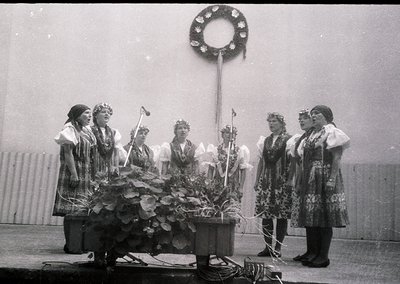 Black-and-white photo of a traditional folk ensemble performing outdoors, likely Eastern European. Seven women in elaborate e...