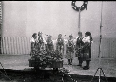 Six women in traditional folk attire perform on a stage, holding floral wreaths and instruments. Mid-20th century Eastern Eur...