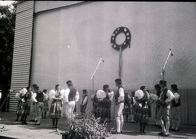 Black-and-white photo of a folk dance ensemble in traditional attire, likely Eastern European, performing outdoors. Men wear ...