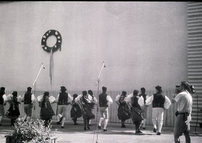 Traditional folk dancers in regional attire perform outdoors, likely in Eastern Europe. Men wear vests, knee-length pants, an...