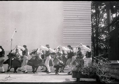 Traditional folk dancers in elaborate costumes perform outdoors, likely in a 1960s–1970s Eastern European setting. Men wear w...