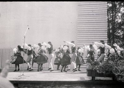 Group of women performing traditional folk dance in coordinated attire—long skirts, blouses, and headscarves—under a micropho...