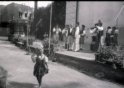 Black-and-white street performance featuring a young dancer in traditional skirt and headpiece, accompanied by a live folk or...