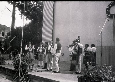 Black-and-white street performance featuring a traditional folk ensemble in 1960s-70s attire: accordion, violin, cello, and p...