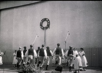 Black-and-white photo of a traditional folk dance performance, likely Eastern European, featuring men in vests, knee-length p...