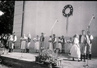 Group performing traditional folk dance on stage, likely Eastern European . Men in vests, women in embroidered dresses and he...