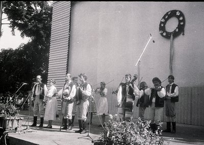 Traditional folk ensemble performing outdoors in 1960s Eastern European attire—men in embroidered vests and women in long ski...