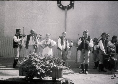Traditional folk ensemble performing in traditional attire, likely Eastern European (1960s-70s). Seven musicians—four playing...