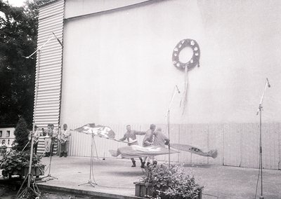 Black-and-white shot of a 1960s-70s stage performance featuring a trapeze act. Four performers mid-air on a suspended trapeze...