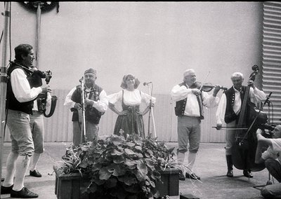 Five musicians perform outdoors in folk attire, likely Eastern European . Violinists, a cellist, accordionist, and vocalist s...