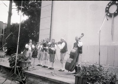 Six musicians perform outdoors on a stage with a corrugated metal backdrop. Instruments include violins, a cello, and a saxop...