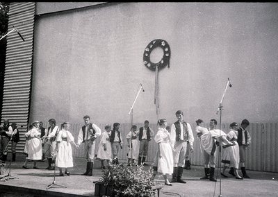 A black-and-white group performance featuring 14 individuals in traditional folk attire, likely Eastern European, performing ...
