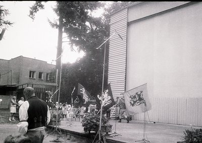 Black-and-white shot of a formal outdoor ceremony, likely 1960s–1980s Eastern Bloc. Stage features floral centerpiece, three ...