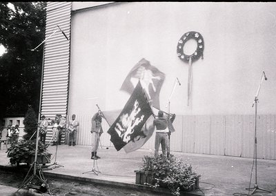 Black-and-white photo of a public ceremony featuring a large banner with a hammer-and-sickle emblem and text in Cyrillic. Sta...