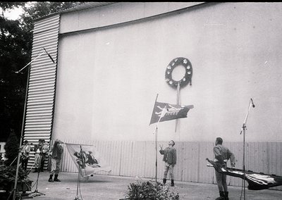 Mid-20th century formal ceremony at a modernist building with clean lines. A wreath and flagpole with a flag bearing a hammer...