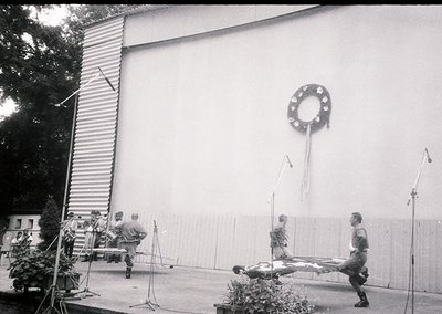 Black-and-white stage setup featuring three performers in coordinated costumes (tunics, knee-high socks, sandals) performing ...