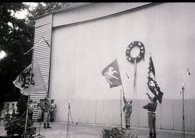 Black-and-white photo of a formal flag-raising ceremony at a large, plain concrete structure. Four individuals in uniform rai...