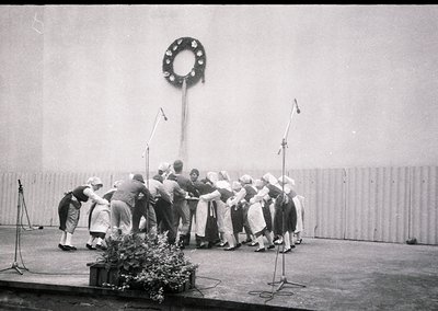 Group of women in traditional folk attire performing a circle dance, likely a Bulgarian *horo*, under a decorative metal ring...