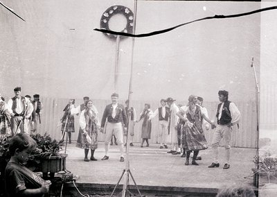 Traditional folk dancers in regional attire performing on an outdoor stage, likely mid-20th century. Men wear vests, hats, an...