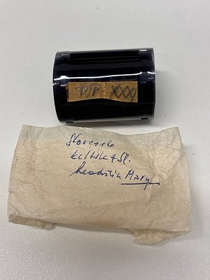 Vintage black cylindrical stamp with embossed "100" and geometric design, resting on aged paper with handwritten note: "Sloan...
