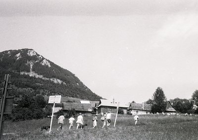 Mid-20th century rural scene: children playing soccer in a grassy field with a wooden fence and mountain backdrop. Simple woo...