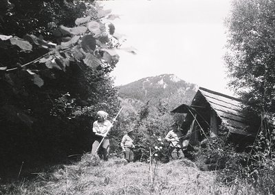 Three individuals in traditional alpine attire gather near a rustic wooden shelter in a forested mountain valley. Mid-20th ce...