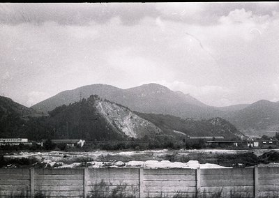 Mid-20th century black-and-white landscape featuring a dam with snow-capped rocks below and forested mountains in background....